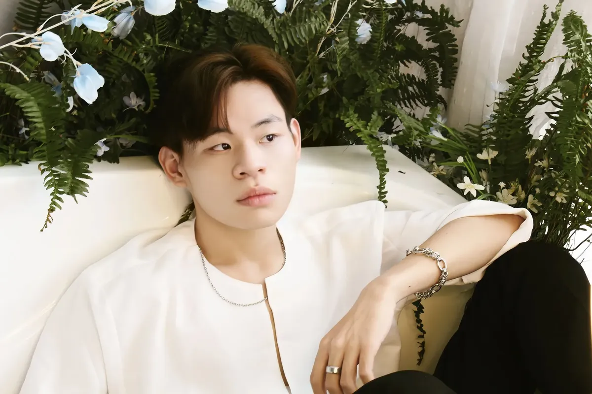 Asian man sitting elegantly on a sofa surrounded by lush greenery. Wearing jewelry.