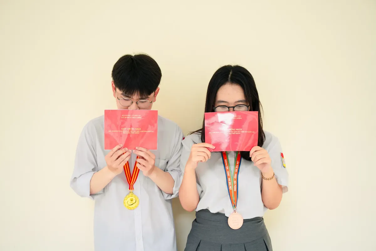 Two Asian teenagers holding diplomas and wearing medals, symbolizing academic success.
