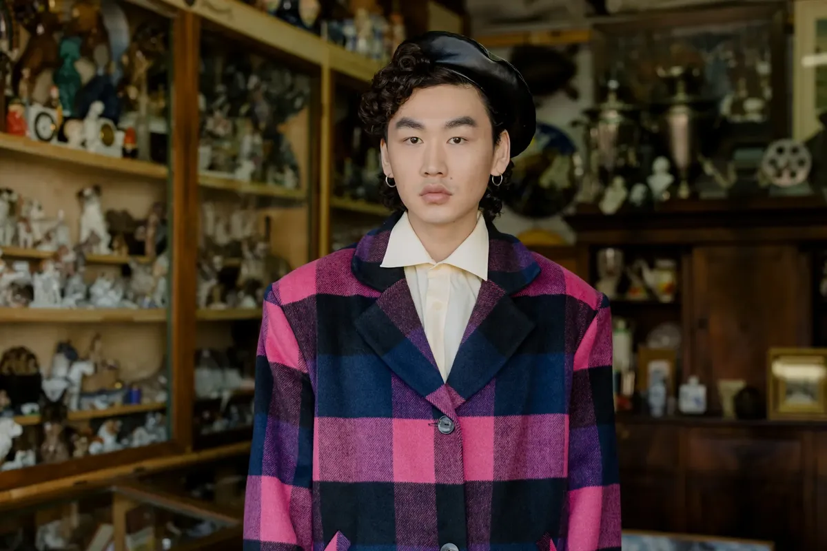 Fashionable man poses indoors wearing a pink checkered coat, showcasing a modern style.