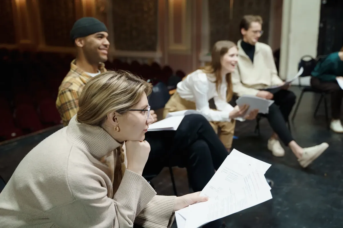 Actors rehearsing a play on stage, holding scripts and discussing roles in a theater setting.
