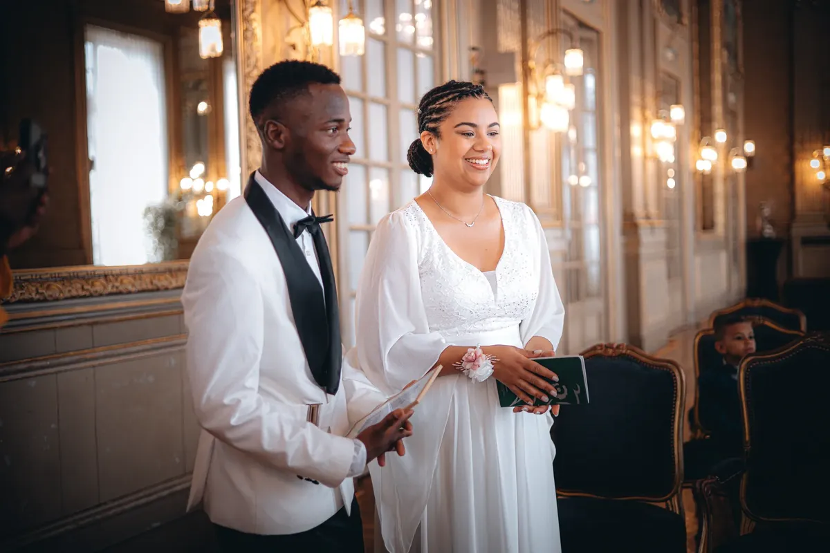 A cheerful couple stands together during their wedding ceremony in a luxurious, ornate hall.