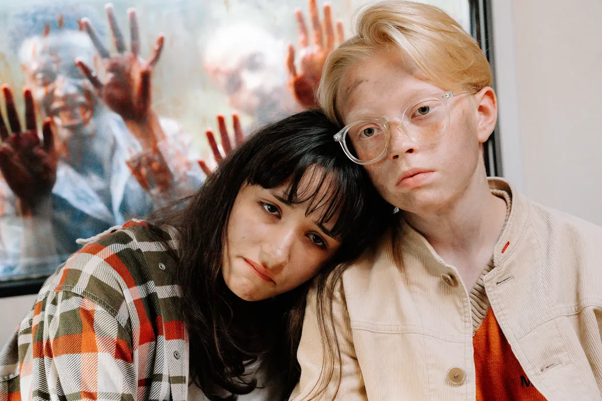 Two people indoors looking worried, with zombies pressing against a window.
