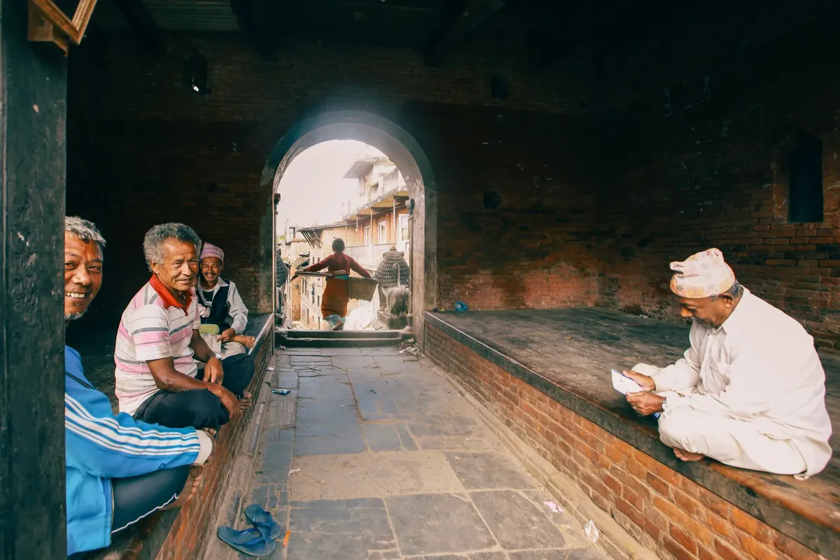 Men gather in a rustic setting with a view of a traditional neighborhood through an archway.